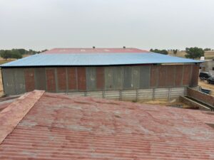 An exterior shot of a large, rectangular warehouse-style building with a red and blue corrugated metal roof. The walls are a mix of corrugated metal in gray and reddish-brown, with numbers painted on some sections. In the foreground, part of a reddish-brown corrugated roof is visible. The background shows a flat, rural landscape with scattered trees under an overcast sky.