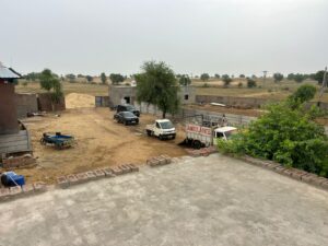 A view of a farmyard with several vehicles parked on a dirt and gravel area. An ambulance-like truck and a white pickup truck are prominent. In the background, there are various small structures, piles of building materials, and an expansive rural landscape with scattered trees under a cloudy sky.