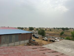 An elevated view showing a section of a farm. In the foreground, part of a red corrugated roof is visible. To the left, a large building with a blue and red roof stands. In the middle ground, several vehicles, including an ambulance-like truck, a white pickup, and a black car, are parked on a dirt area. The background extends into dry, open land with sparse trees under a light grey, overcast sky.