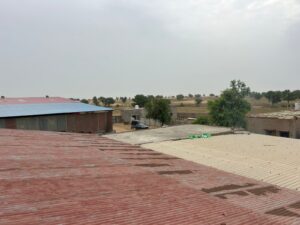 A view over rooftops towards a rural landscape. In the midground, a black car, a white pickup truck, and a white ambulance-like vehicle are parked on a dirt lot. Several small buildings are visible behind them, surrounded by dry, open land with sparse trees under a cloudy sky.